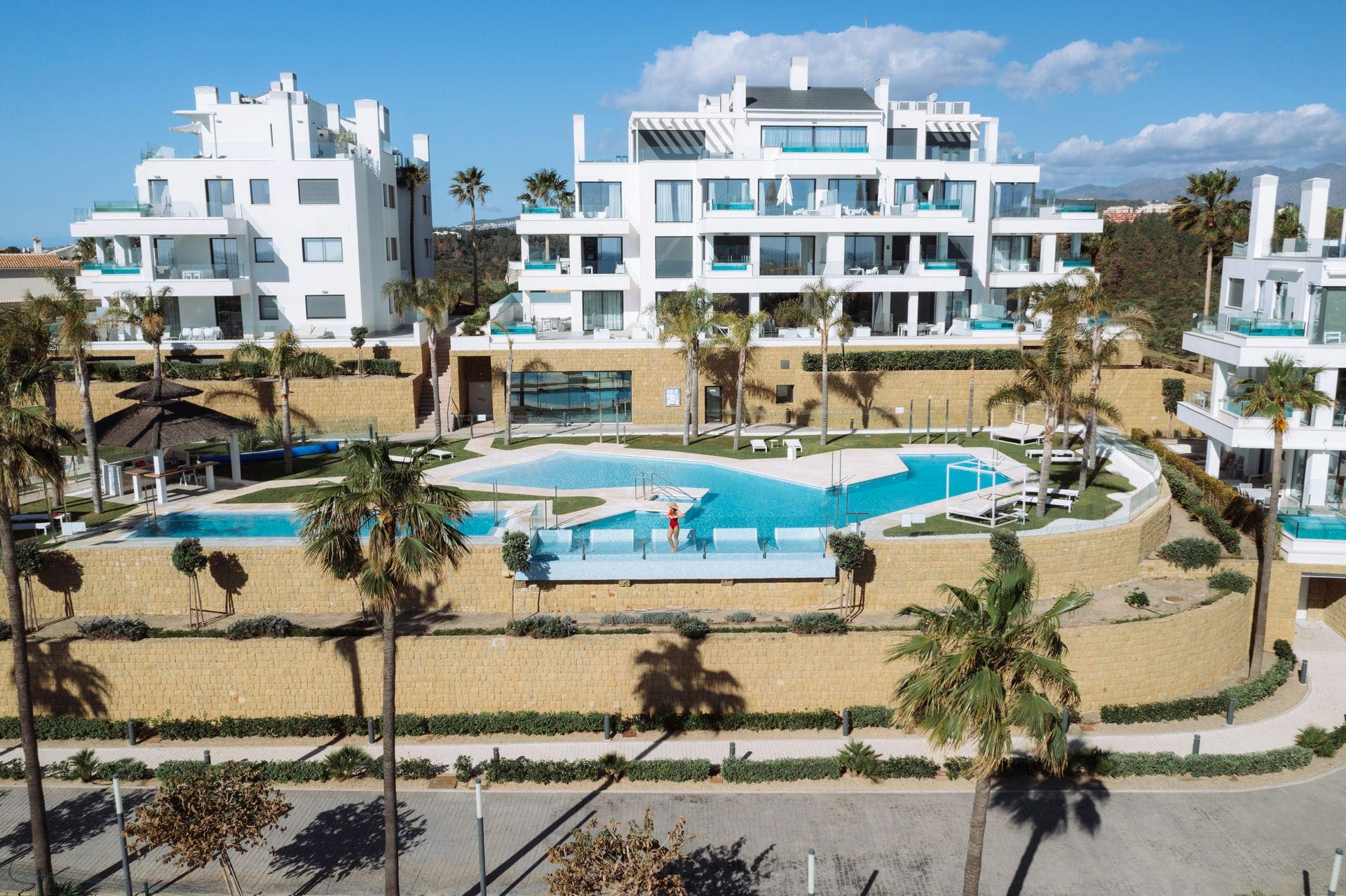An IDILIQ development on the Costa del Sol: white terraced apartment blocks above a run of pools and sandstone retaining walls, palms throughout, the sierra behind them.